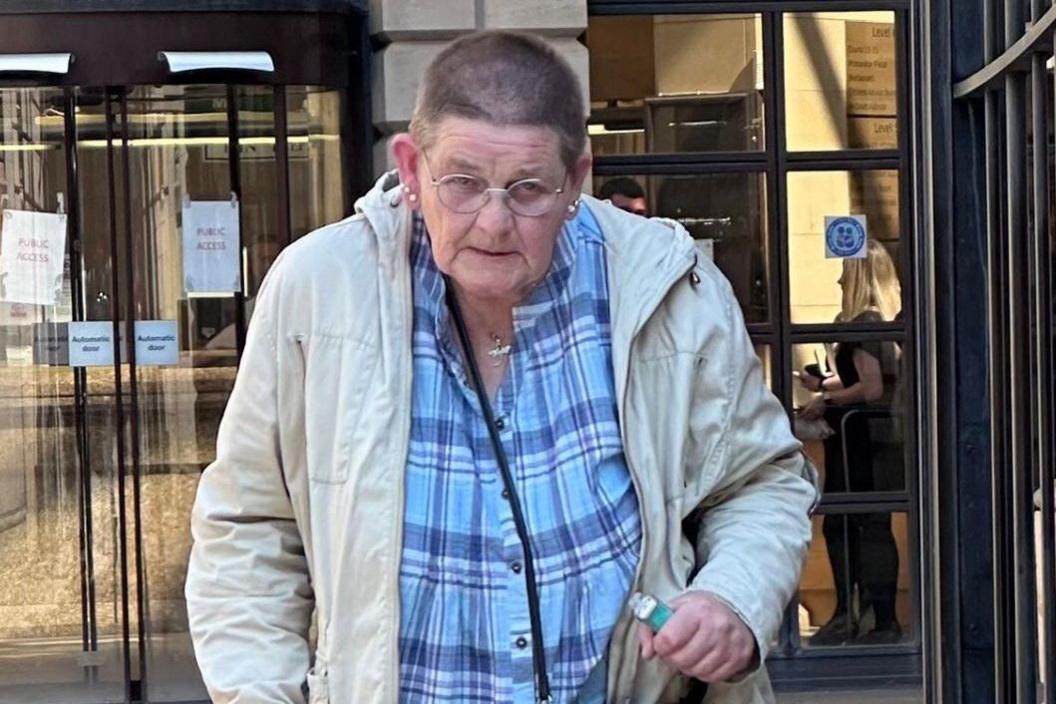 A woman with short cropped hair and glasses, walking with the aid of a stick outside a court