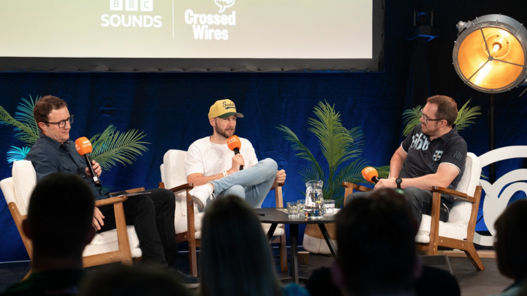 Three men sit on white chairs in front of an audience. They are holding black microphones with orange covers.