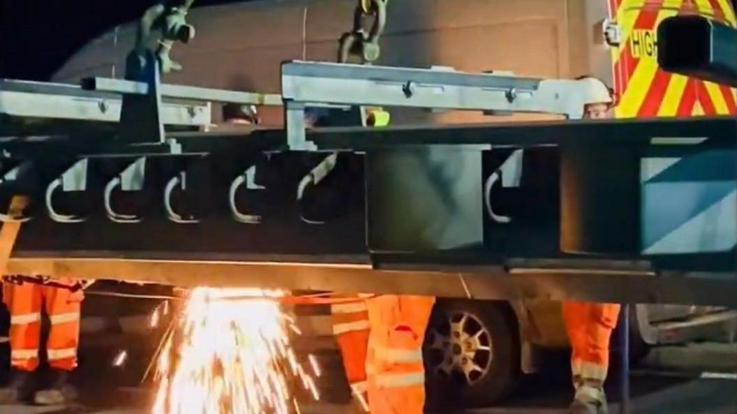 A new bridge joint being put into place by works in orange high-vis outfits. There are sparks from metal work being carried out. 