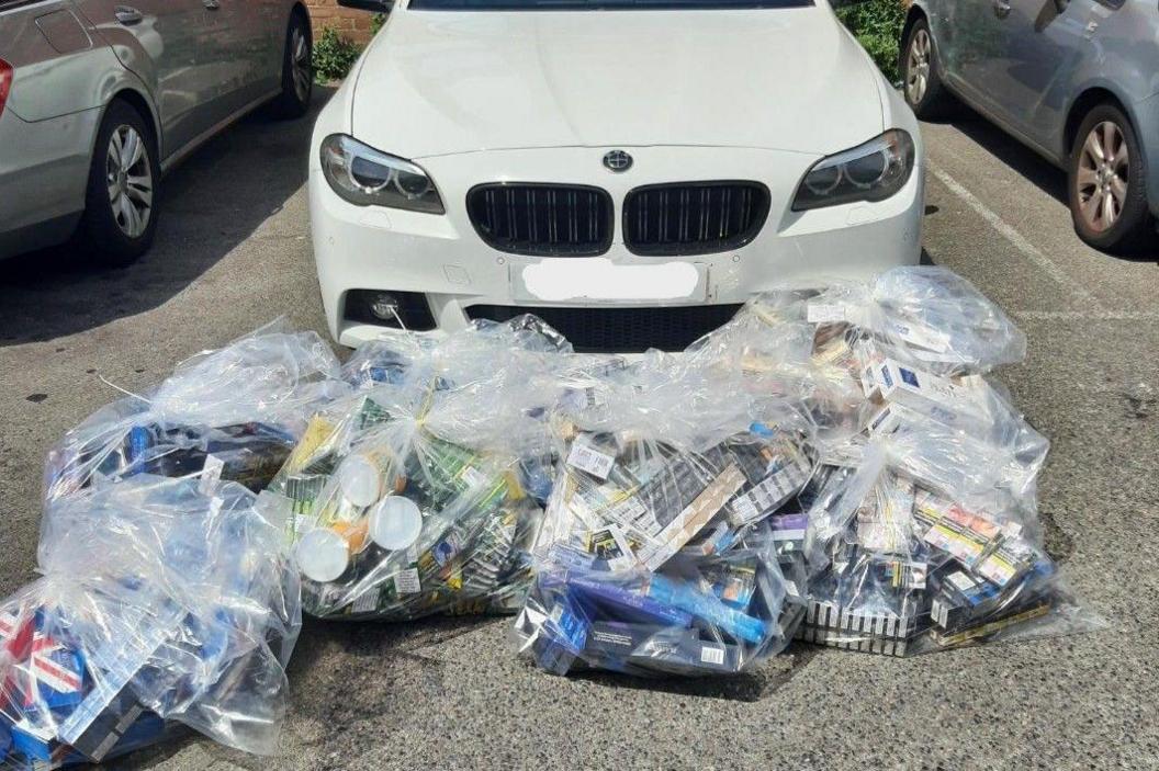 Bags of tobacco boxes are piled up in transparent bags in front of a white BMW.
