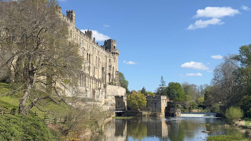 The high side of a castle on a hill overlooking a waterway with trees and grass on both banks