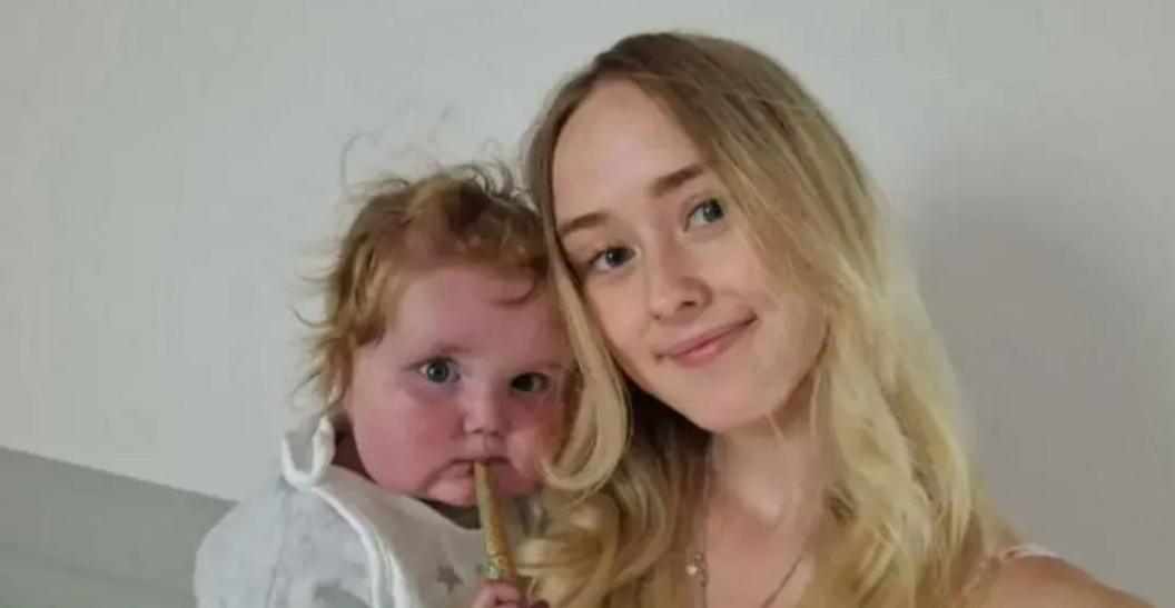 A woman with long blonde hair holding a baby with curly blonde hair, a white top and a breadstick in his mouth