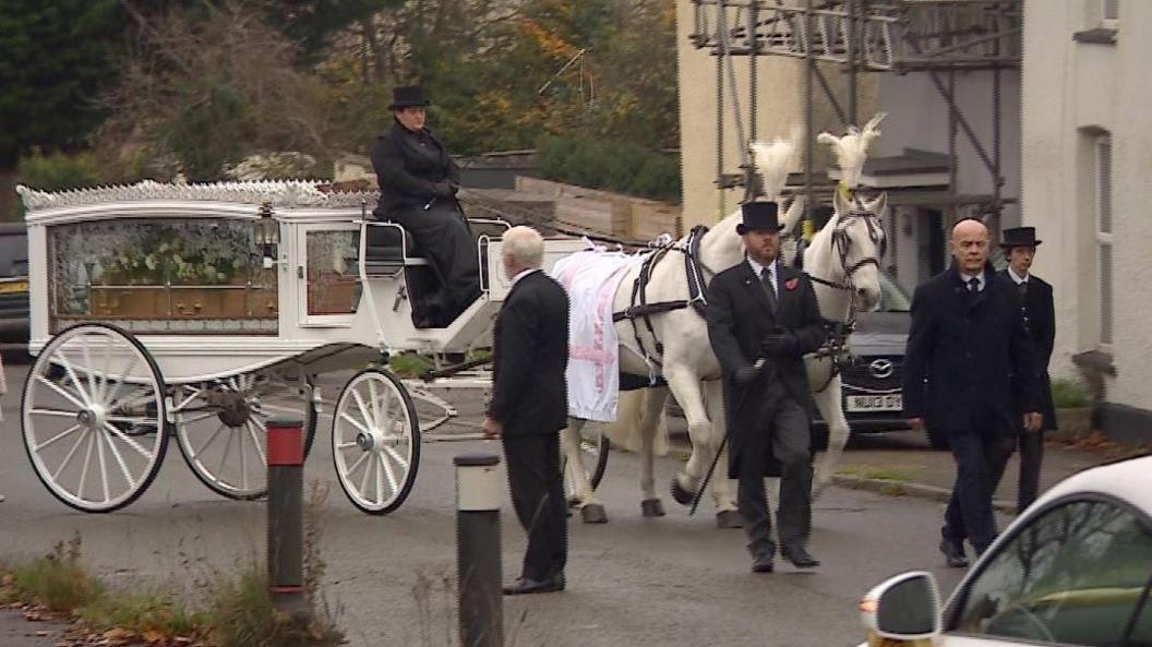 A funeral procession with two white horses pulling a carriage with a coffin inside. The coffin has white flowers on top. Four men in black suits are walking in front of the horses.