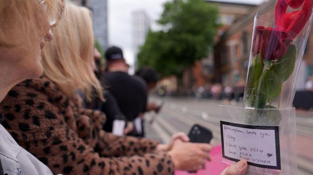 A woman with strawberry blonde hair and glasses smiles towards an empty road. She is holding a red rose and a red heart inside a plastic wrapper. A note on the front reads "Birmingham loves you, I love you Ozzy, Love Eliza Finn O'Shea"