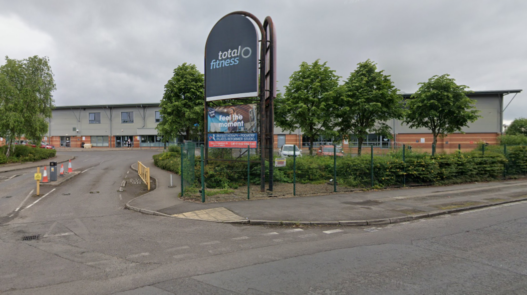 A large sign reading 'Total Fitness' - the site where the proposed music academy will be built. The sign sits in front of a large warehouse on an industrial site.