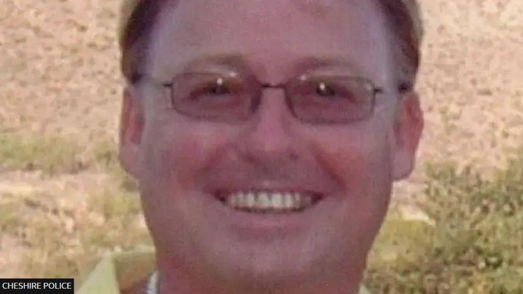 A man is smiling while looking at the camera. He has short brown hair and is wearing glasses.