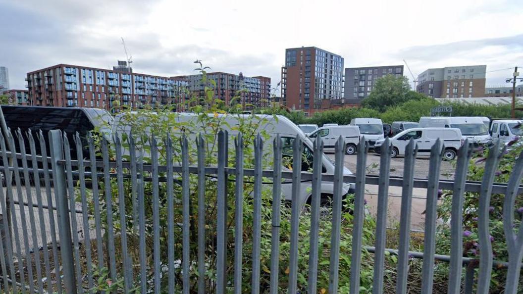 A google street view image of the vacant plot on Upper Wharf Street in Salford. White vans can be seen parked all over the site behind grey metal fencing, with apartment blocks seen looming in the background. 