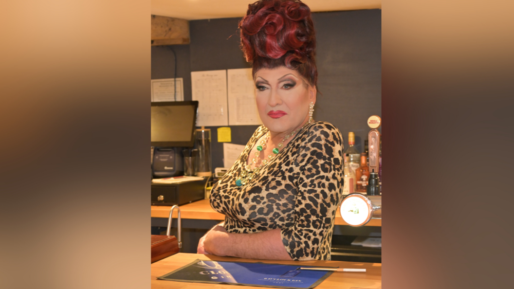 A man in drag, he wears a leopard print top, necklace and leans on a bar in a pub setting. He has typical drag make-up, thick red lipstick and strong eyebrows, and a red beehive.