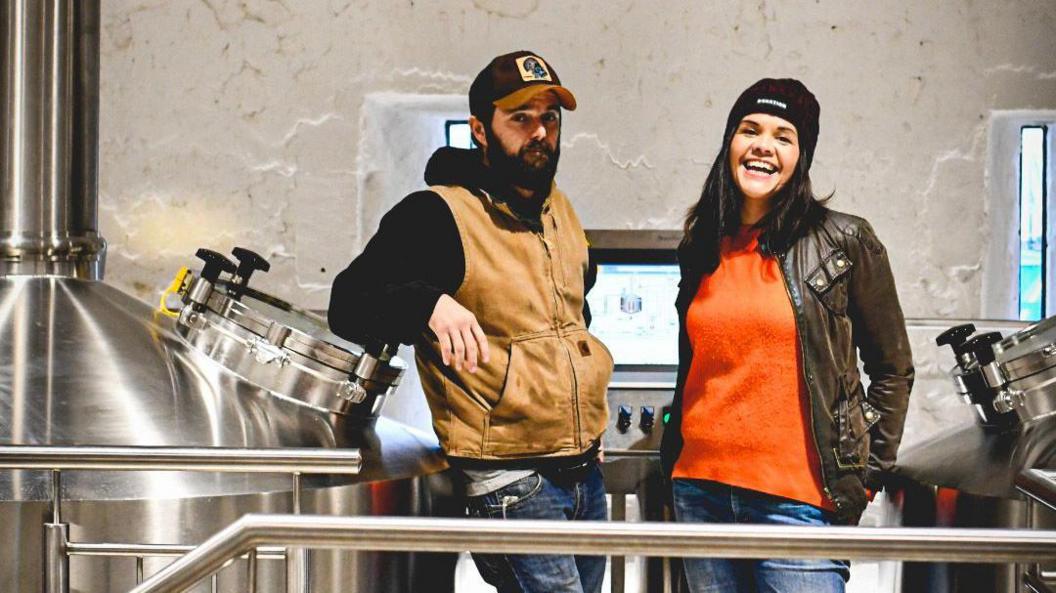 Miranda Hudson and Derek Bates stand in a brewery, with a computer screen behind them and stainless steel tanks either side. Derek is on the left and has a dark beard. He is wearing a black hoodie, brown gilet, blue jeans and a black and brown baseball cap. He has a neutral expression. Miranda is wearing a black beanie hat, orange jumper, a wax cotton or leather jacket and blue jeans. She has shoulder-length dark hair and is laughing.