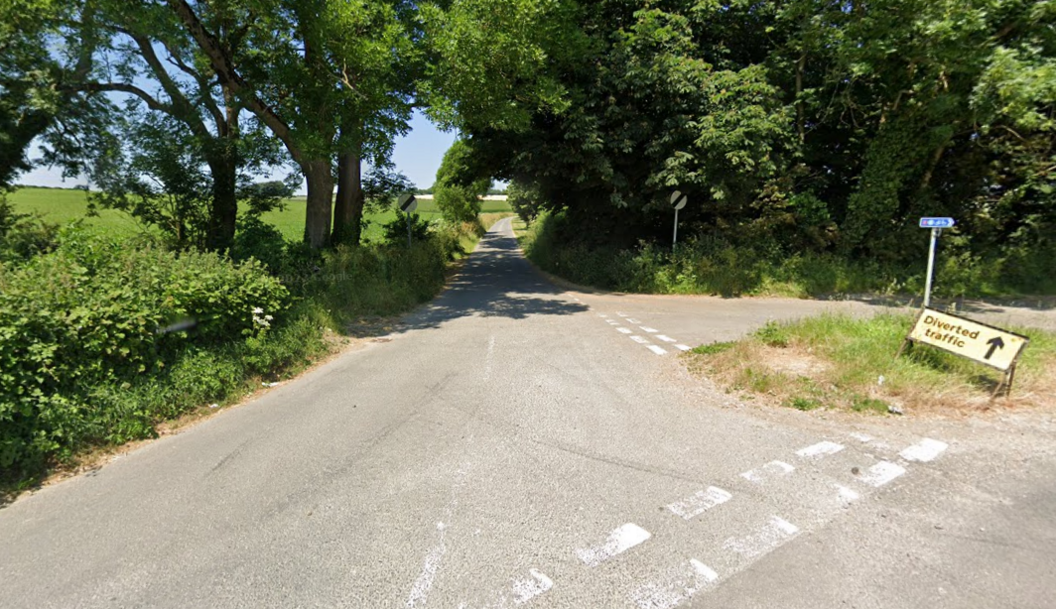 Bats Lane, close to the junction with Mallards Green, with faded road markings on the right hand side of Bats Lane and a diverted traffic sign pointing towards Mallards Green. It is a rural lane, with large trees and bushes surrounding the road 