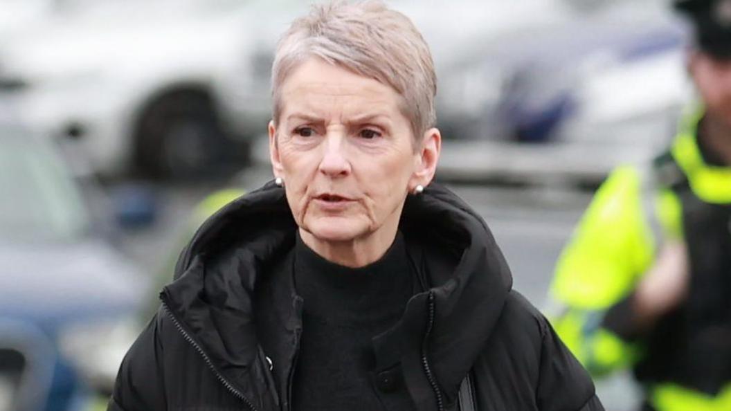 Lady Eleanor Donaldson arrives at Newry Crown Court. She wears a black puffer coat and black top. She has short grey hair. There are cars parked behind her. There is a police officer in a yellow high vis jacket behind her.