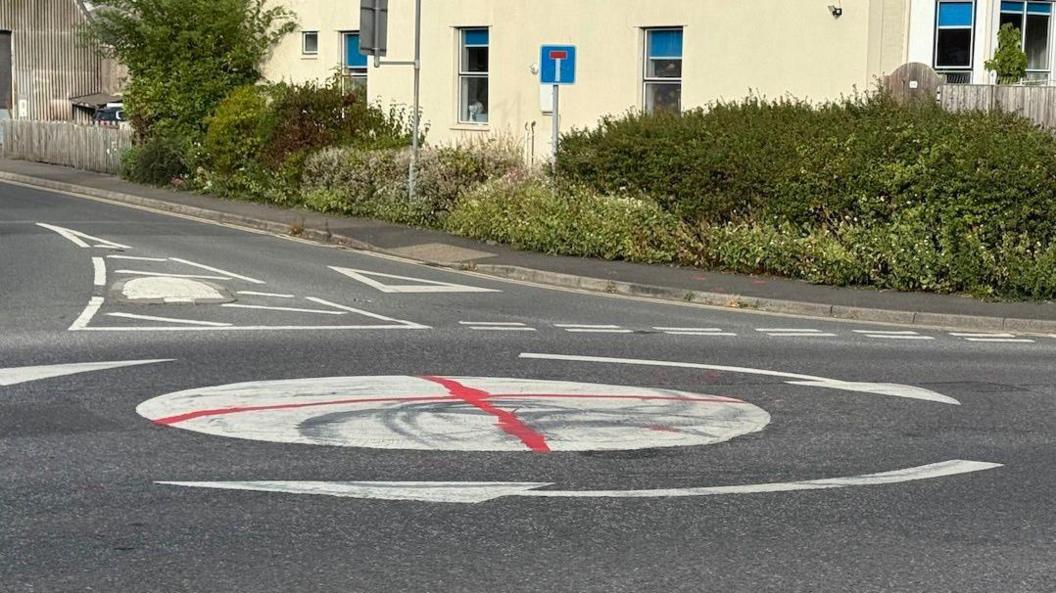 A mini white roundabout in Yatton with a spray painted red cross on it in a residential area, with what appears to be a large cream rendered block of flats or a large house beside the junction.