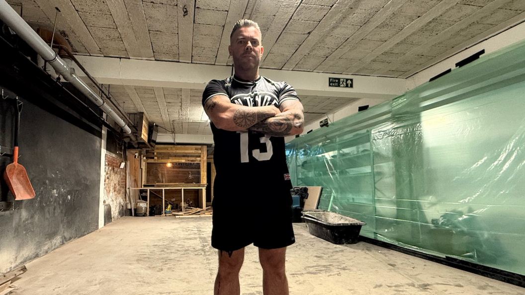 Garry Bell, standing with crossed arms and wearing shorts and a T-shirt, inside Contender Gym which is dirty and looks like it's in the process of being rebuilt, with no proper floor covering, plastic sheeting covering bits of gym kit, raw wood frames and what looks like a bath for mixing concrete in the background.