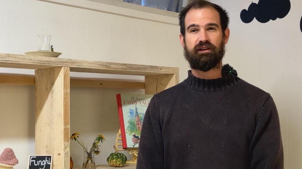 A man in a dark jumper with dark hair and a beard standing next to some shelves