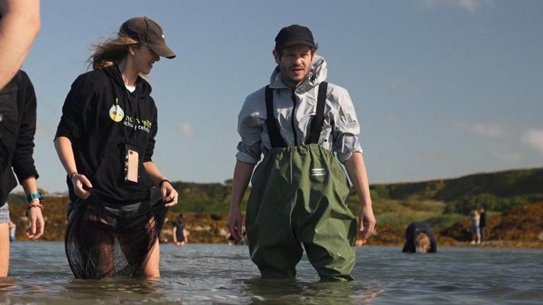 Iwan Rheon standing thigh deep in water wearing green waders