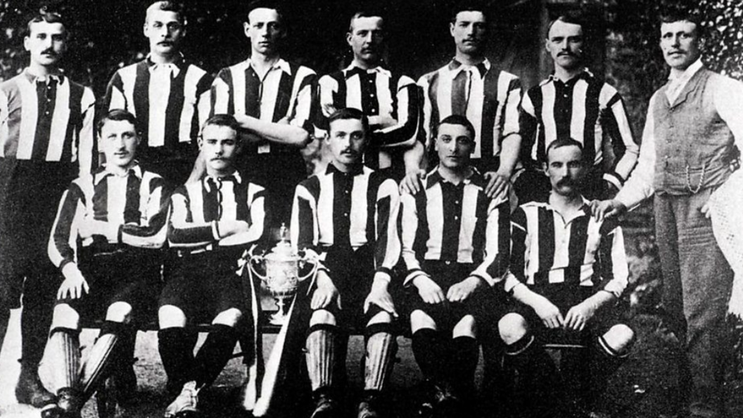 A black and white image of footballers in striped kit, seated in two rows, with a man in a suit to the righthand side of the image.

On the lower row, an ornate cup is perched on the edge of a chair between two of the footballers.