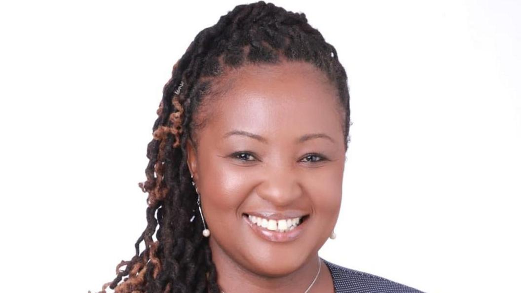 Ms Kilonzi is seen from the shoulders up against a white background. She has brown eyes and is smiling. Her hair is in a half up, half down do and has jewel drop earrings in. 