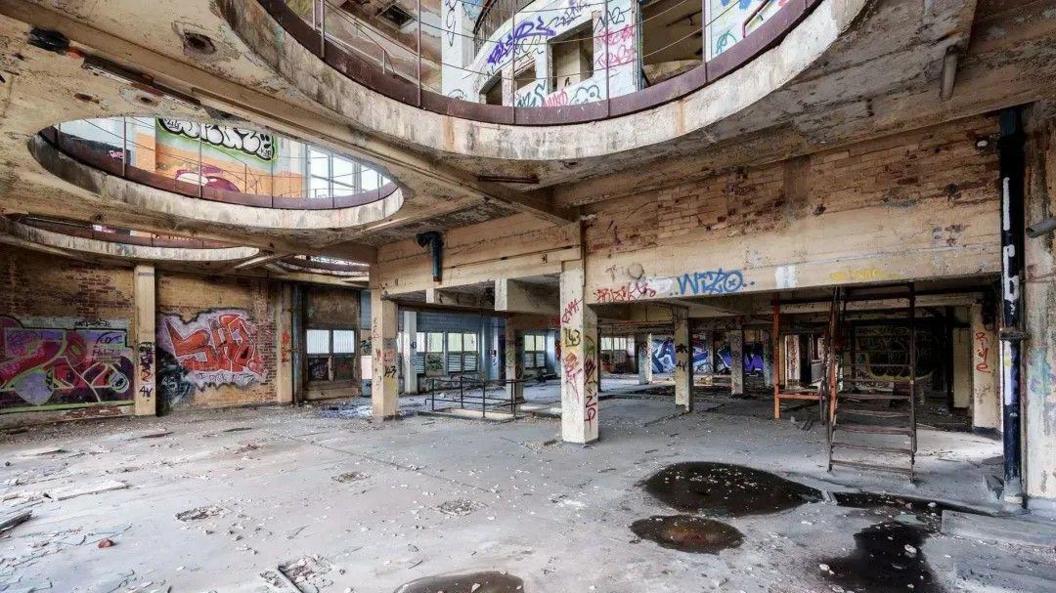 Inside a derelict building. There are tiles falling off the walls and dust from the degrading building. There are lots of graffiti tags and other spray paint around the walls. The ceiling above has two large circular "holes" with safety barriers around the ledges bove.