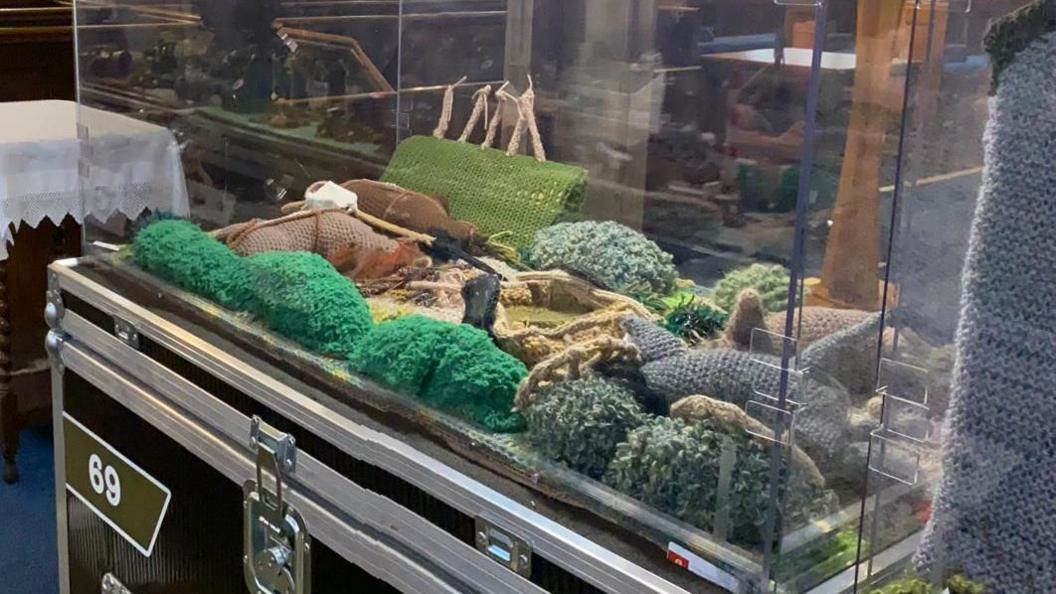 A knitted scene from the D-Day landings, using various shades of green wool, is inside a clear box.