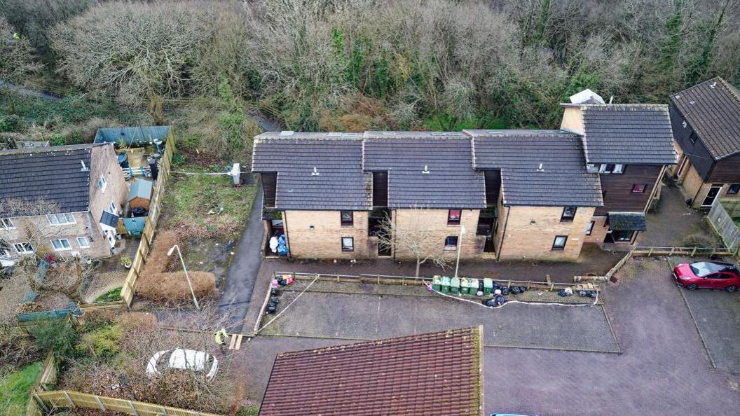 An aerial view of three blocks of flats with police tape outside. In front of the flats is a car park with several bins and two cars parked. Another property is visible to the left of the frame, and otherwise the scene is surrounded by green trees.