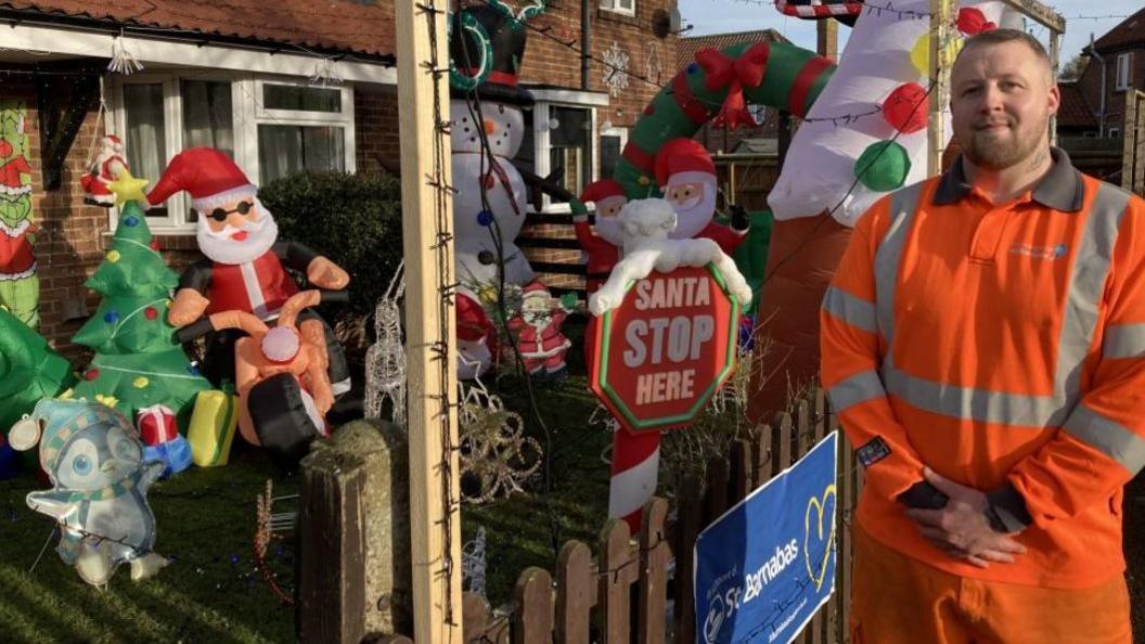 A man is standing on the right wearing orange high vis with Anglian Water written on the front. He has short brown hair and a short brown beard and is smiling at the camera. He is stood next to the front of his house, which has a blow-up Christmas tree, snowmen, Santa, and a sign saying 'Santa stop here'. It is daylight but there are several lights on the display and a St Barnabas sign on the fence.