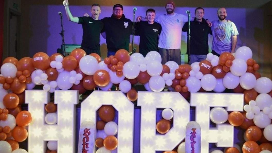 Six men on a stage in front of balloons and microphones