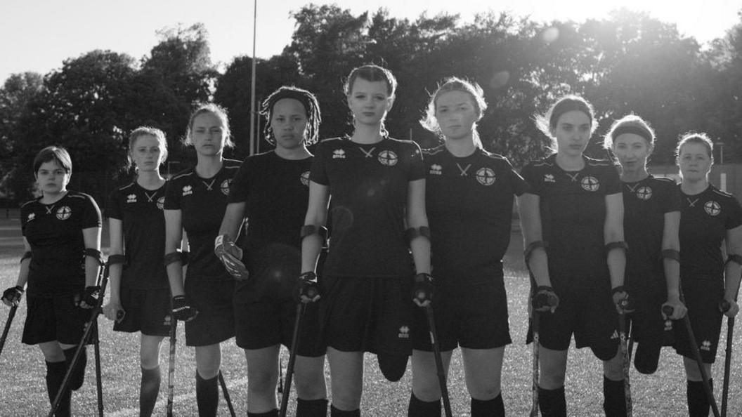 A black and white picture showing nine members of England Women's amputee football team, dressed in black shorts, t-shirts and football socks, standing on a football pitch. Eight of the women have crutches and five can be seen to have had a leg amputated.