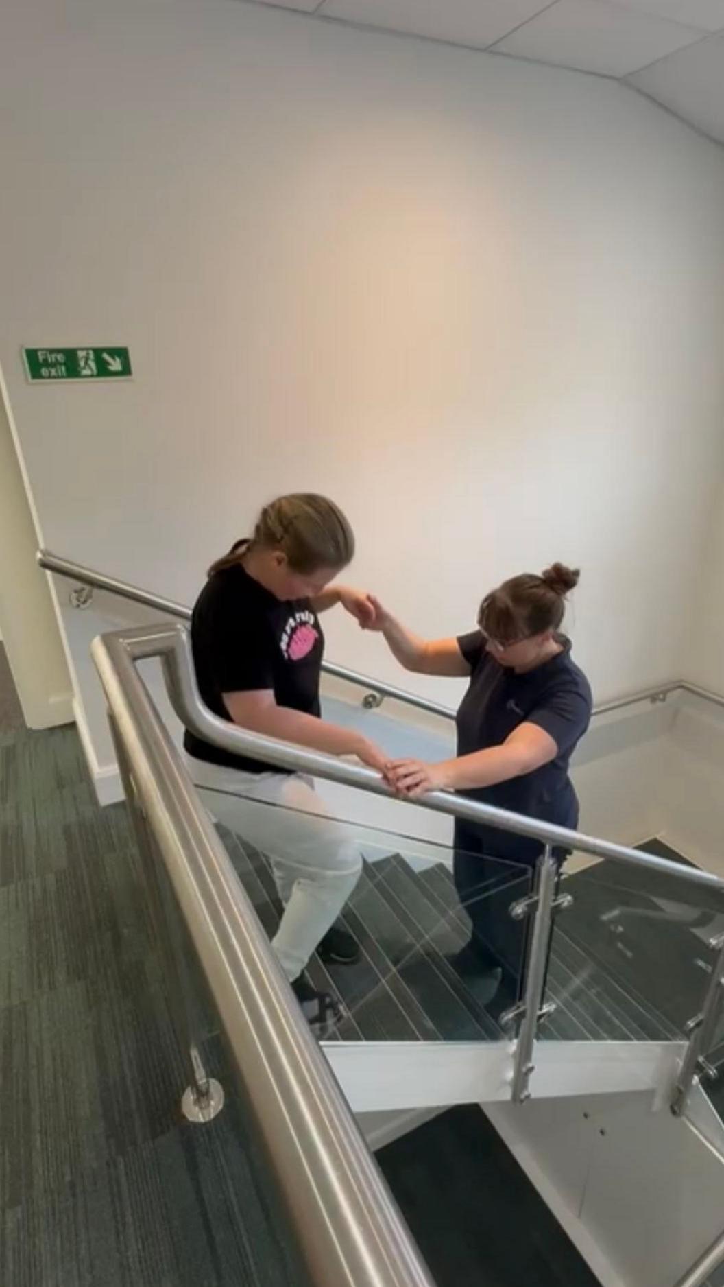 The image is taken in a stairwell with white walls, grey carpet and chrome handrails with glass inserts. It shows a woman with light brown hair, tied back and wearing a black t-shirt with a pink logo and light coloured joggers at the top of the stairs, she has her right hand on the rail and is holding the hand of another woman who is a few steps below her facing upwards. They are both looking down towards the steps. The woman who is facing upwards is wearing black trousers and a black t-shirt and has dark brown hair in a bun with a fringe and is wearing brown framed glasses.