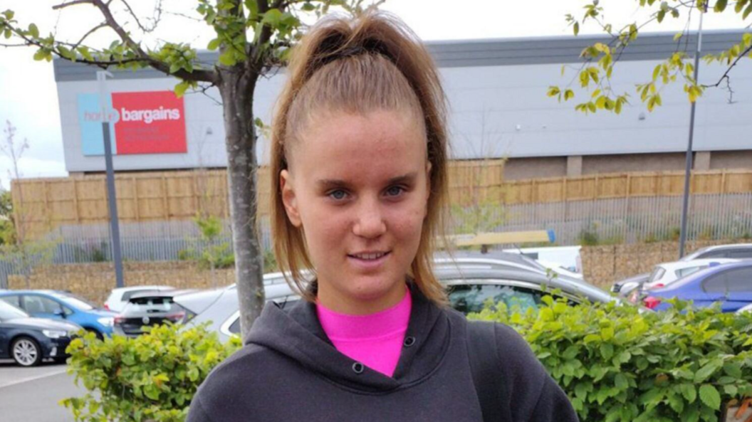 Holly standing in a car park. She has long light brown hair.