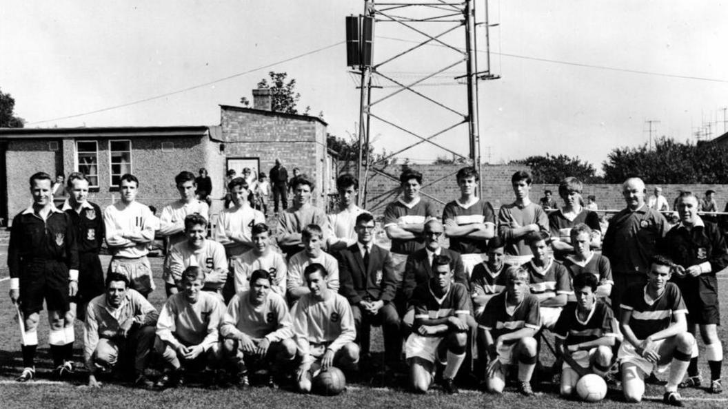 A black and white photo of footballers lined up in three rows with match officials at Cambridge United's Abbey Stadium in about the 1950s