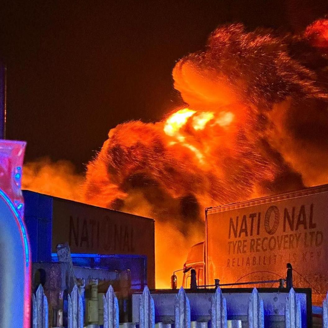 Plumes of smoke, lit orange, rise high in the sky. There are two HGVs in front of the flames. One of them says National Tyre Recovery Ltd. The spikes of a fence are visible at the bottom of the image.