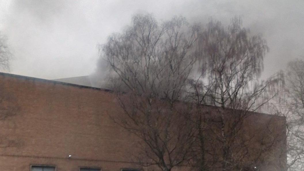 Smoke billowing into the sky from the top of a large brick building. The building is pictured from the street and has no windows visible. Trees can be seen growing next to it.