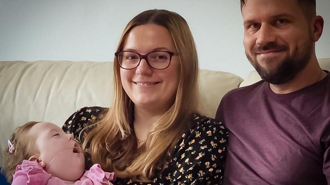 Baby Robyn in a pink jumper. She has long blond hair, her eyes are closed and she has a breathing tube in her nose. Mum also has light hair a printed blouse. She is hold Robyn and smiling. Dad Daniel is in a T-shirt and is snuggled in to his wife on their couch