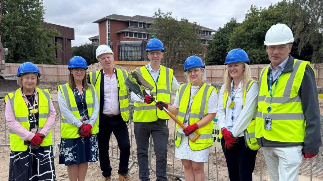 People stading arranged in a semi-circle- two woman, two men, two women and  a man- all wearing blue helmets and green high-visibility jackets- standing at the construction site with one of the ladies from the right holding a yellow spade.