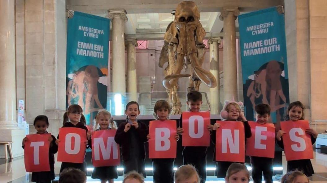 Children stand in front of a mammoth replica in a museum holding individual red paper spelling out 'Tom Bones' in white lettering.