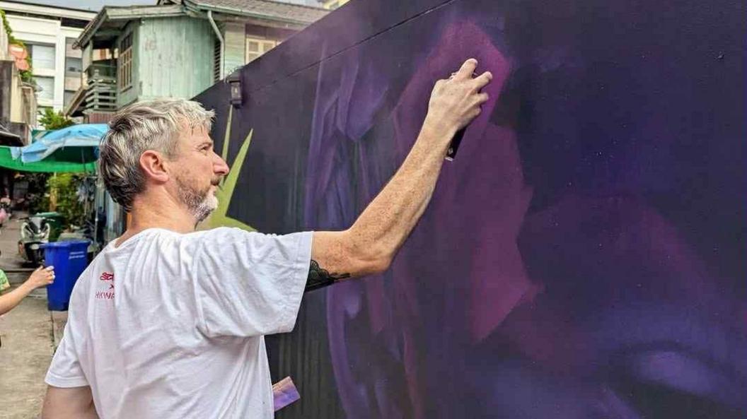 A man with grey hair in a white t-shirt is painting a mural on a dark wall. The mural is a mix of blue and purple and resembles the side of a persons head. The wall is located in an alley in Bangkok which can be partially seen in the background