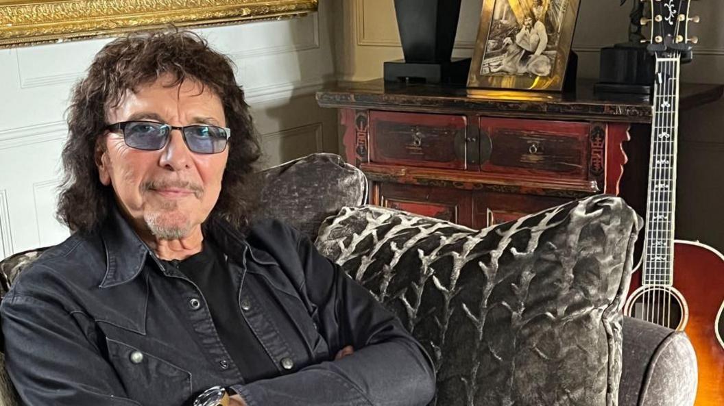 A man with black hair, dark glasses and a black top on a grey sofa with a guitar behind him