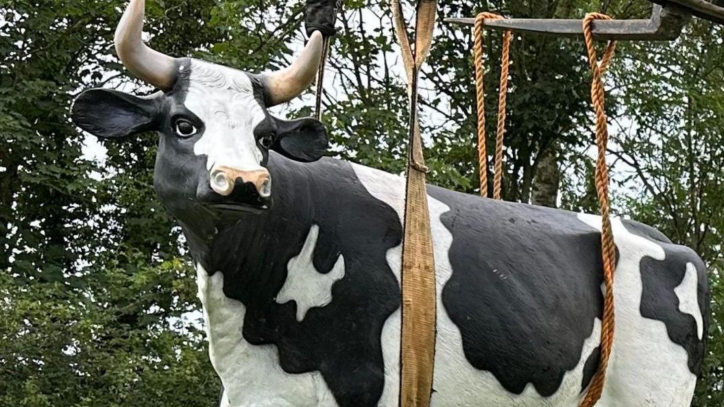 A life-sized plastic cow, with horns, sat in a harness. Some trees are in the background.