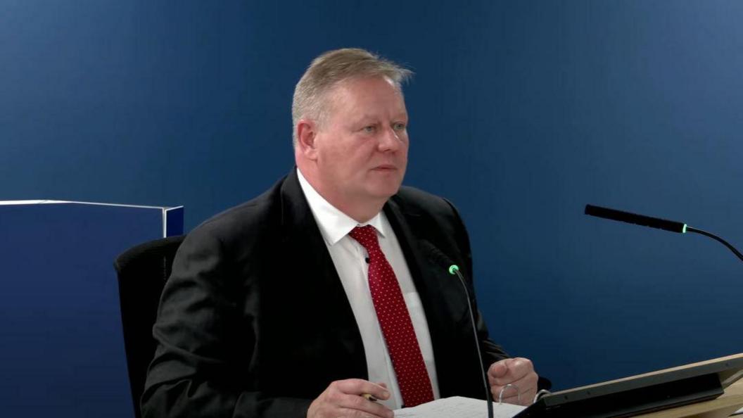 A man, with short grey hair, sitting at a desk. He is wearing a white shirt, red tie and black suit. There are two microphones on the table in front of him. 