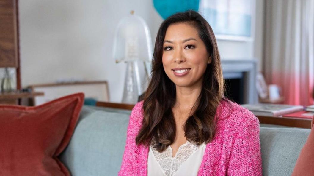 Vivien Wong wearing a white lace camisole and bright pink wool cardigan sitting on a grey sofa in a plush home