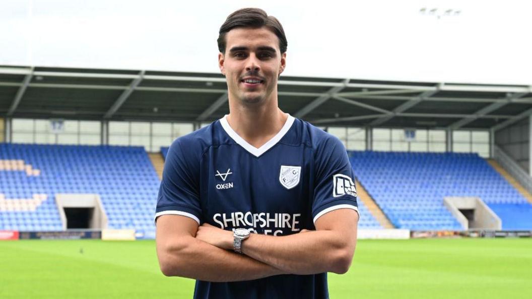 Isaac Fletcher standing with arms folded in dark blue Shrewsbury Town shirt with a stand at Croud Meadow in the background