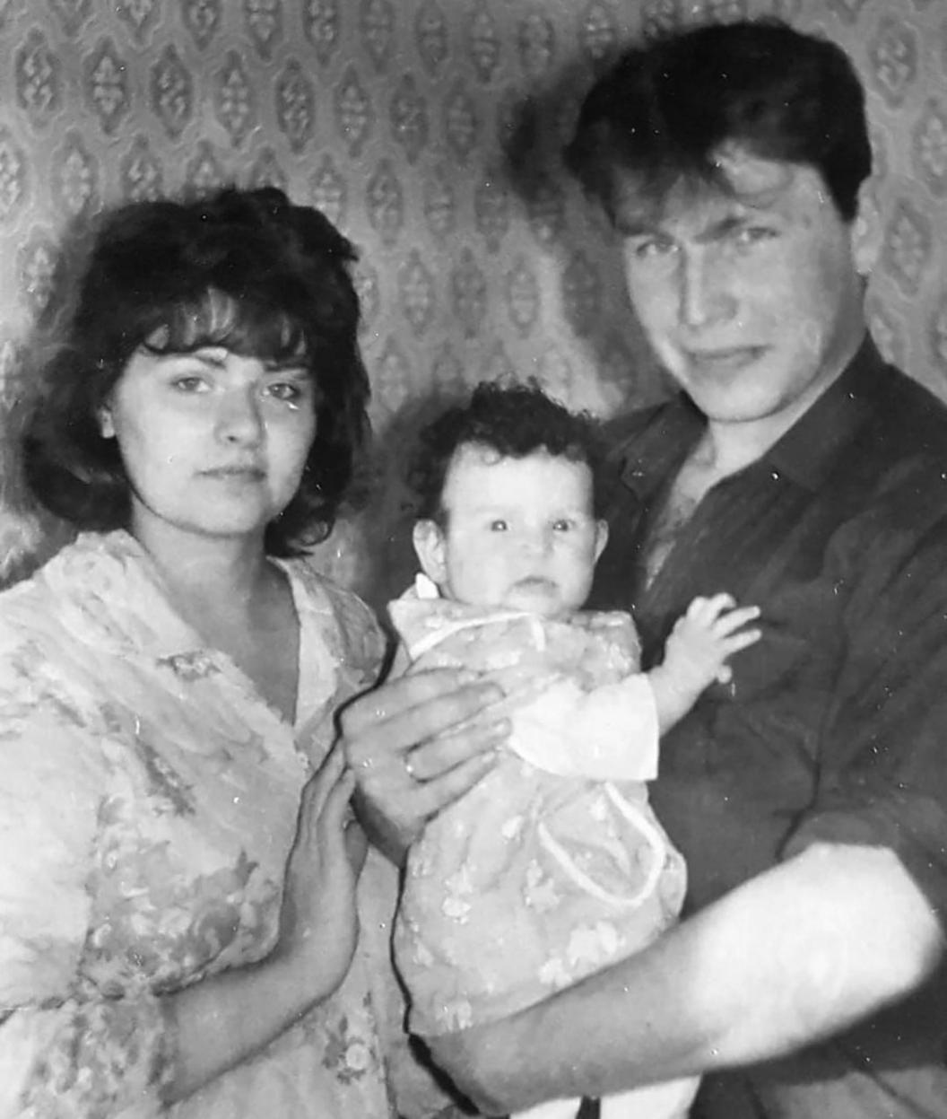 Black and white photo of Iryna and Serhiy with baby Katya. Serhiy is wearing a dark shirt and holding the baby, who has dark hair and is looking at the camera. Iryna is wearing a floral blouse.
