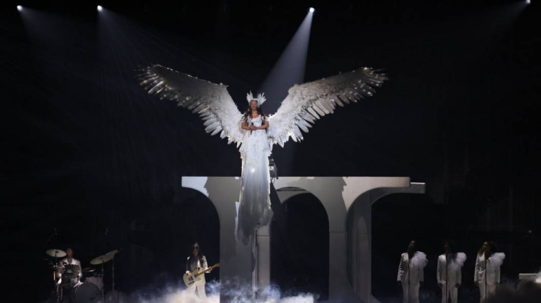 Jade Thirlwall performs during the 2025 BRIT Awards ceremony at The O2 arena in London. She wears huge white wings and a winged headband. She is dressed all in white and is lifted above white columns while a band also dressed in white plays below.