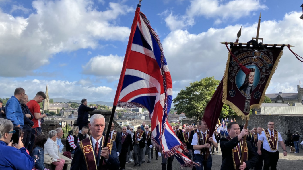Members of the Apprentice Boys wearing sashes and one carrying a Union flag, with spectators watching on and city buildings visible in the background