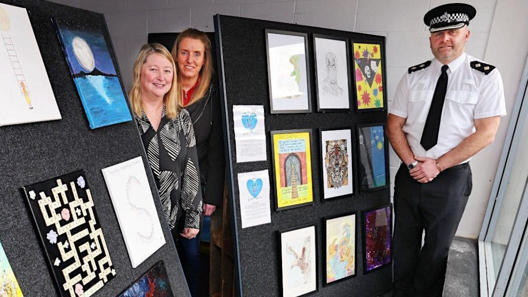 Amanda Murphy and Kelly Smith stand in between two displays of artwork with Insp Alan McKeon standing on the right. They are all smiling. A range of art can be seen including a colourful image of a lion and a train track leading into a tunnel.