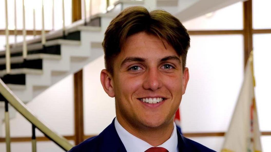 George Finch looks at the camera. He has floppy short brown hair with a centre parting, and wears a suit with a red tie.
