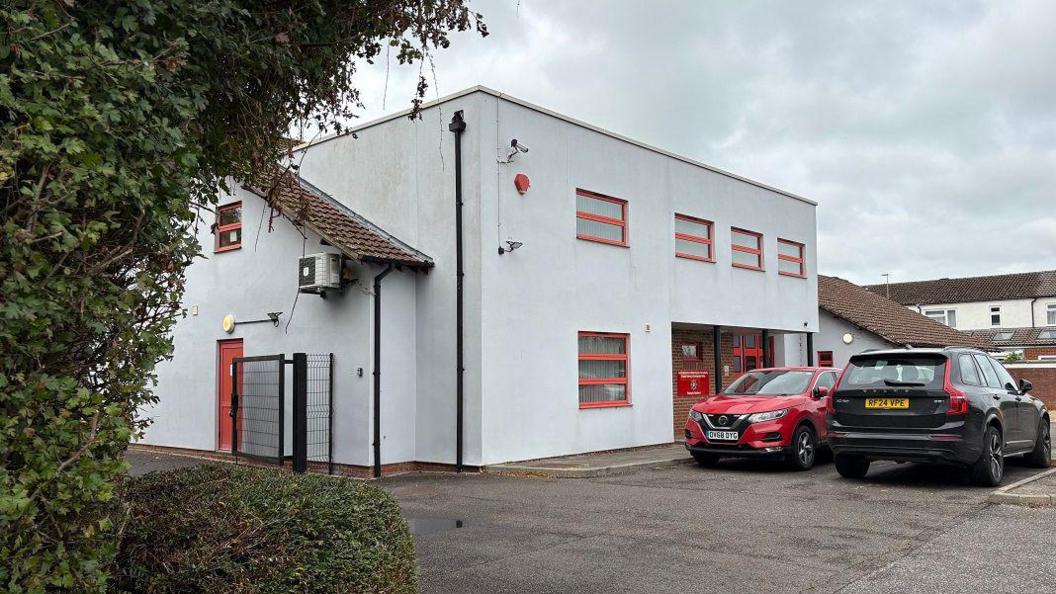 Two-storey white fire station with red-framed windows. Red doors are visible at the front of the building. There are two cars parked at the front. There is a hedge to the left of the picture.
