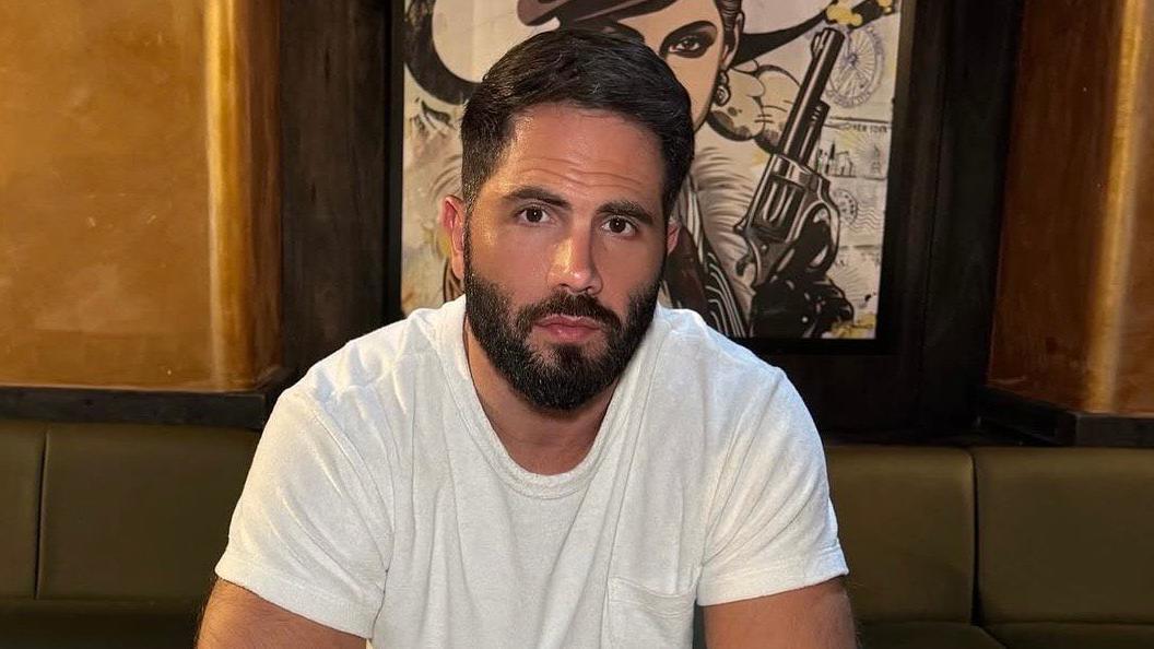 Hass, who has brown eyes, black hair and a beard, looks into the camera. He is wearing a white t-shirt and sitting on brown leather seating, with a painting of a woman holding a smoking gun behind him.