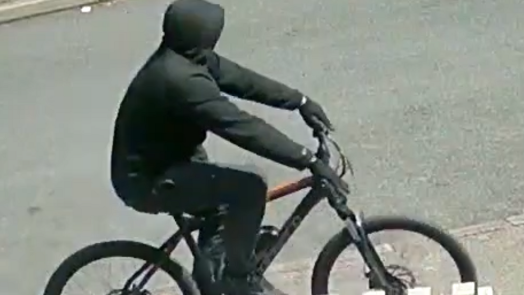 Another image of a man dressed completely in black, riding a black bicycle in front of a white fence