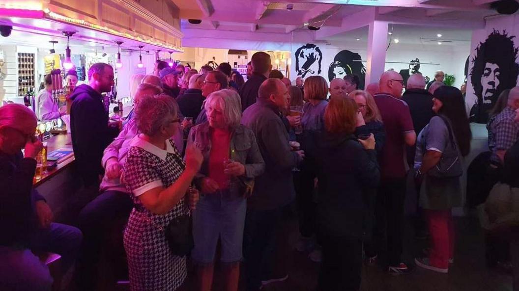 A bar in a room, full of people chatting and drinking, with pictures of Jimi Hendrix, David Bowie and Amy Winehouse painted on the wall.